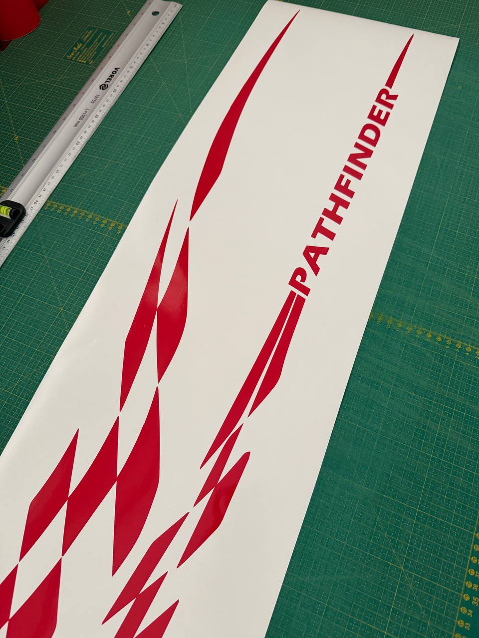 A long white vinyl decal sheet featuring red geometric chevron patterns and the word 'PATHFINDER' in red, laid diagonally on a green cutting mat with a ruler visible in the corner.